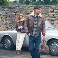 A Lake District scene depicting a man and woman wearing Lakeland Leather clothing including jackets, footwear and bags. A classic British sports car is in the background, parked in front of a stone wall.