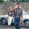 A Lake District scene depicting a man and woman wearing Lakeland Leather clothing including jackets, footwear and bags. A classic British sports car is in the background, parked in front of a stone wall.